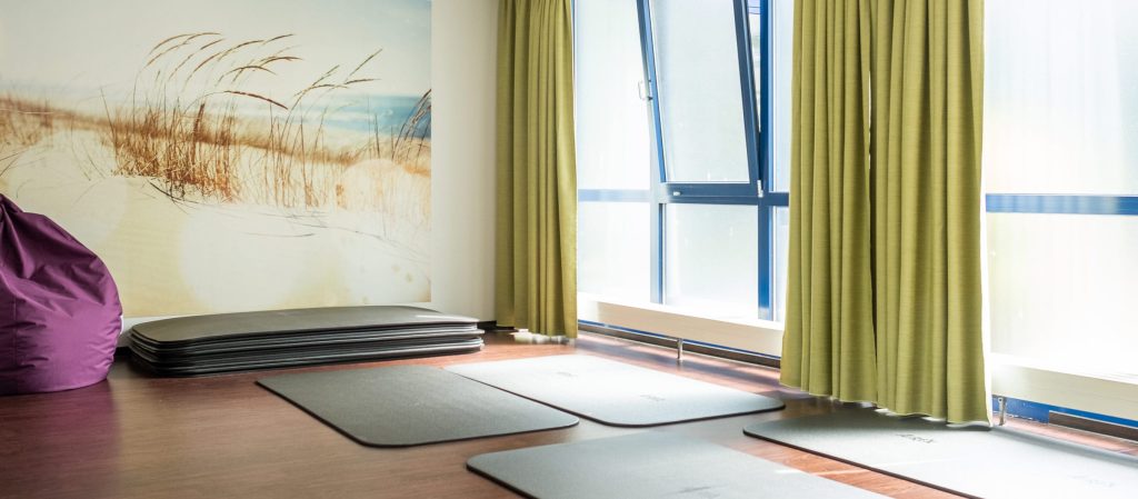 Ein heller Gymnastikraum in der Ostseeklinik Grömitz mit großen bodentiefen Fenstern rechts. Auf dem dunklen Holzboden liegen mehrere graue Trainingsmatten bereit, im Hintergrund stapeln sich weitere Matten. Die Wand links ist mit einer großformatigen Fototapete gestaltet, die eine Dünenlandschaft an der Ostsee zeigt. Ein violetter Sitzsack setzt einen farbigen Akzent, während olivgrüne Vorhänge die großen Fenster einrahmen.