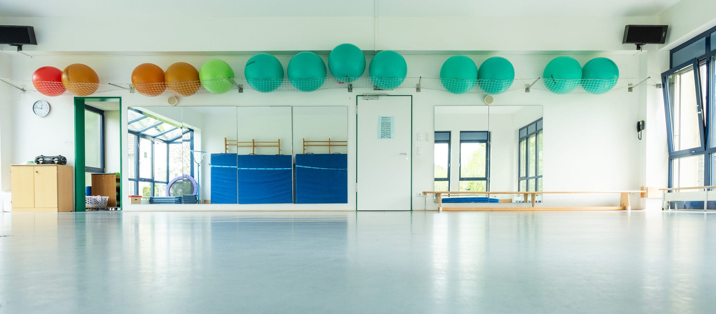 Heller, lichtdurchfluteter Gymnastikraum der Ostseeklinik Grömitz mit einer großen Spiegelwand, blauen Turnmatten und einer Sprossenwand. An der Wand lagern in einem Netz verschiedene bunte Gymnastikbälle in Rot, Orange, Grün und Türkis; der Boden ist spiegelnd sauber und bietet viel Platz für therapeutische Übungen.