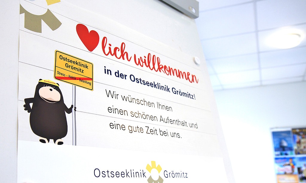 Bei der Anreise: Schild mit der Aufschrift „Herzlich willkommen in der Ostseeklinik Grömitz! Wir wünschen Ihnen einen schönen Aufenthalt und eine gute Zeit bei uns.“ Neben dem Text ist eine Illustration eines Ortsschildes zu sehen, auf dem Stress, Hektik und Belastung durchgestrichen sind.