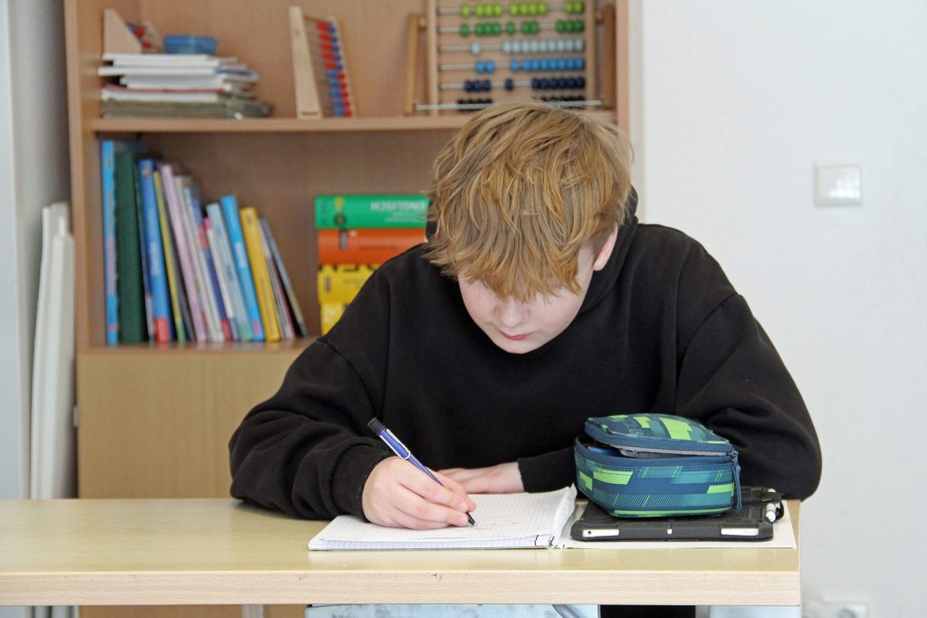 Ein Junge mit rötlich-blonden Haaren sitzt konzentriert an einem Schultisch der Ostseeklinik Grömitz und schreibt in ein Heft. Im Hintergrund ist ein Bücherregal mit Lernmaterialien und einem Abakus zu sehen.