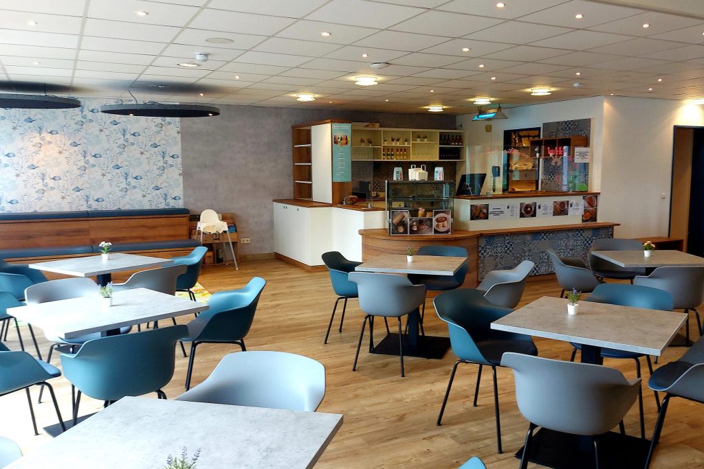 Heller, modern gestaltete Cafeteria der Ostseeklinik Grömitz mit hellgrauen Tischen, blauen Schalenstühlen und einer Service-Theke im Hintergrund.