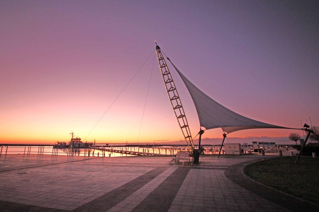 Stimmungsvolle Abenddämmerung an der Ostsee in Grömitz. Die Silhouette der Seebrücke und die moderne Segel-Konstruktion am Strand werden vom warmen Licht des Sonnenuntergangs eingerahmt.