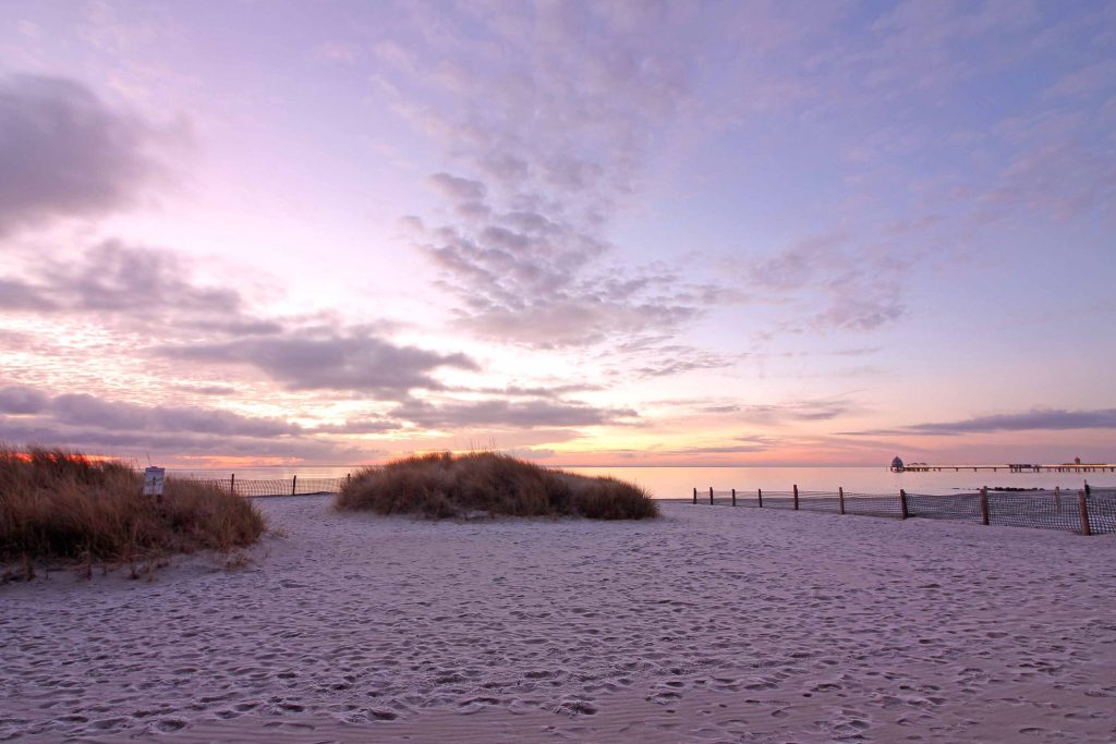 Weite Dünenlandschaft am Ostseestrand von Grömitz bei Sonnenuntergang. Der feine Sand im Vordergrund wird von sanftem, lila-rosafarbenem Abendlicht beleuchtet. Im Hintergrund ist die Silhouette der Seebrücke über dem ruhigen Meer unter einem leicht bewölkten Pastellhimmel zu sehen.