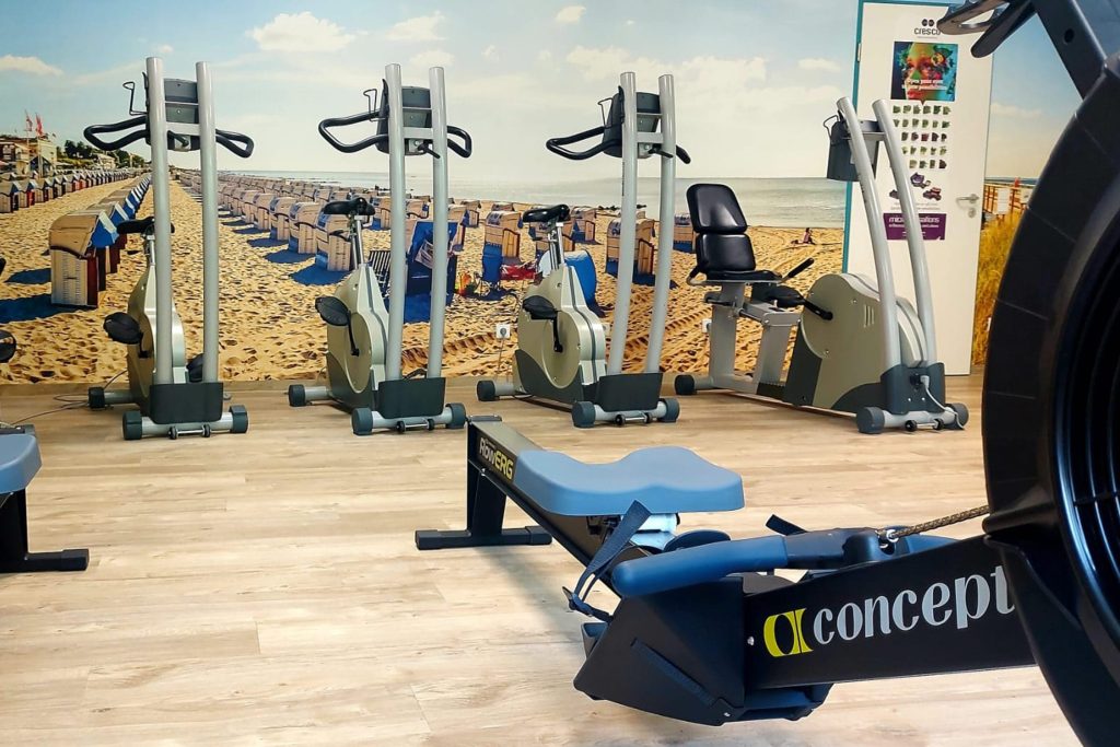 Innenansicht eines Fitnessraums der Ostseeklinik Grömitz. Im Vordergrund steht ein blaues Fitness-Rudergerät. Im Hintergrund befinden sich mehrere Fahrradergometer vor einer großflächigen Fototapete, die den Grömitzer Sandstrand mit Strandkörben zeigt.