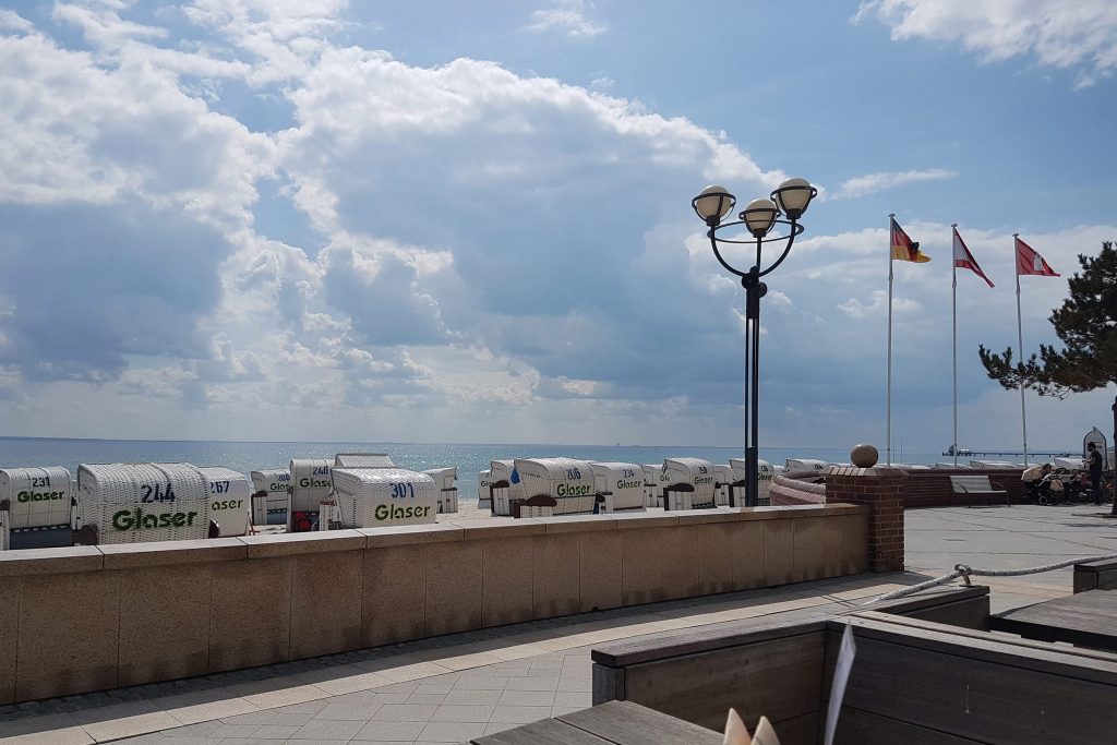 Die sonnige Strandpromenade von Grömitz an der Ostsee nahe der Ostseeklinik. Im Vordergrund sind mehrere weiße Strandkörbe ordentlich nebeneinander aufgereiht. Eine klassische, dreiflammige Straßenlaterne und drei wehende Flaggen säumen den gepflasterten Weg. Der Blick führt über eine niedrige Mauer direkt auf das ruhige Meer unter einem weiten, leicht bewölkten blauen Himmel.