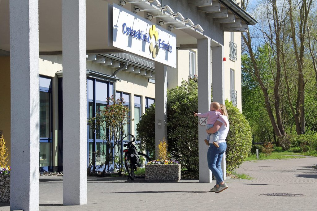 Ein sonniges Foto des Haupteingangs der Ostseeklinik Grömitz. Über dem überdachten Eingangsbereich mit weißen Säulen hängt ein Schild mit dem Kliniknamen und Logo. Im Vordergrund steht eine Mutter in Jeans und weißem Oberteil, die ihr Kind trägt, das in Richtung des Klinikeingangs zeigt. 