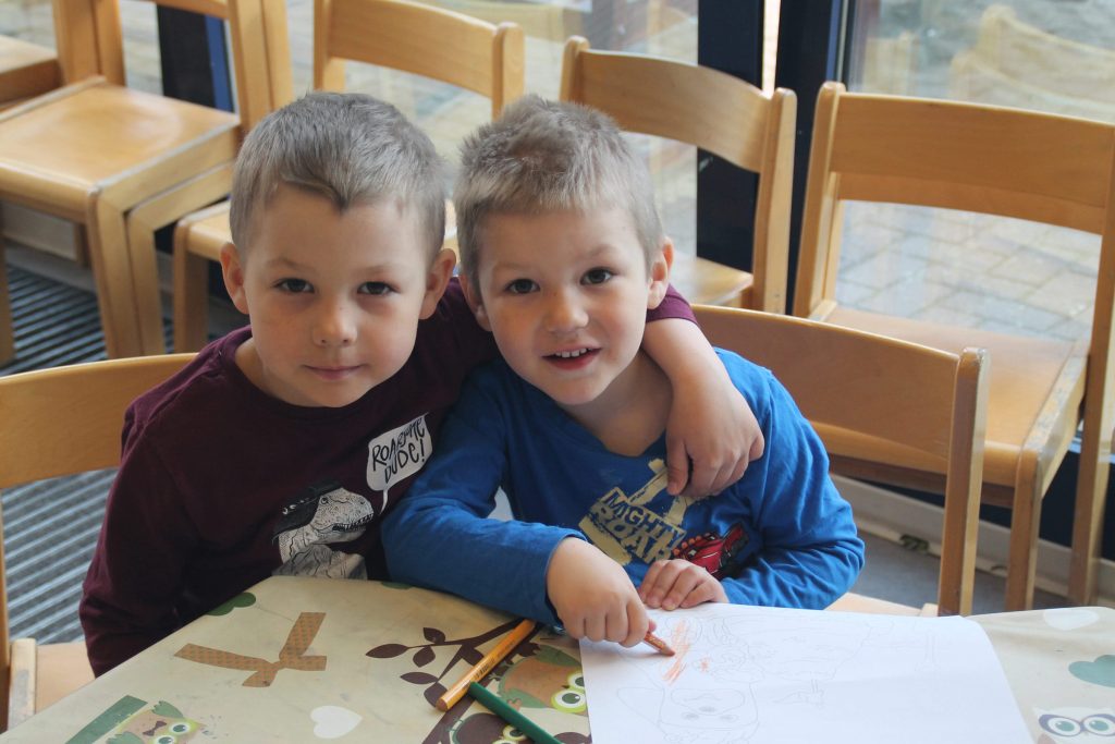 Zwei kleine Jungen sitzen Arm in Arm lächelnd an einem Basteltisch in der hellen Kindertagesbetreuung der Ostseeklinik Grömitz. Im Hintergrund sind kindgerechte Holzstühle und eine große Fensterfront zu sehen.