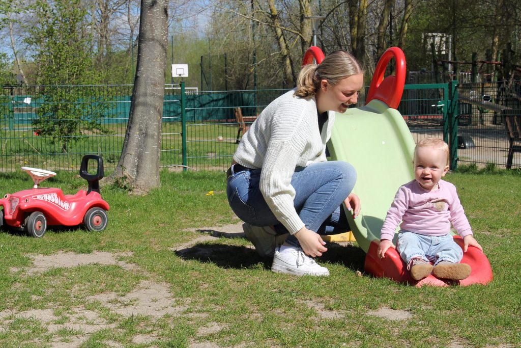 Eine junge Mutter in einem hellen Strickpullover und Jeans kniet lächelnd auf einer Wiese im Außenbereich der Ostseeklinik Grömitz neben ihrem Kind, das gerade eine kleine grün-rote Rutsche hinunterrutscht. Im Hintergrund sind ein rotes Bobbycar, Bäume und ein umzäunter Spielplatzbereich bei sonnigem Wetter zu sehen.