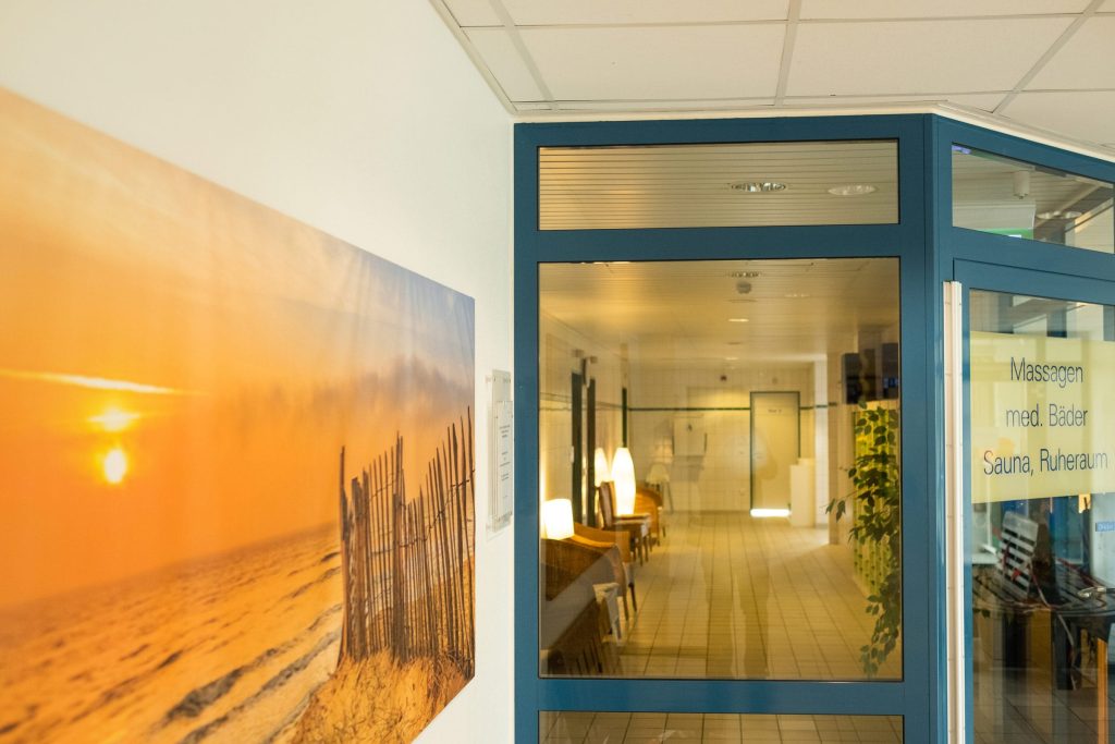 Hell gestalteter Innenbereich der Ostseeklinik Grömitz mit Blick durch eine Glastür in einen Flur mit Sitzgelegenheiten, links an der Wand hängt ein großformatiges Bild eines Sonnenuntergangs am Ostseestrand.