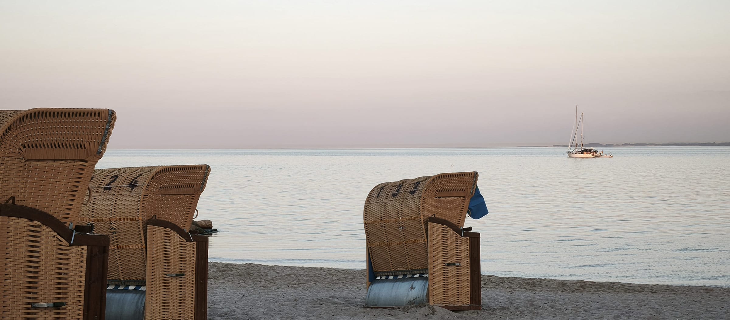 Zwei Strandkörbe im weichen Sand am Strand von Grömitz direkt vor der Ostseeklinik Grömitz mit Blick auf die ruhige Ostsee bei Sonnenuntergang und einem Segelboot in der Ferne.
