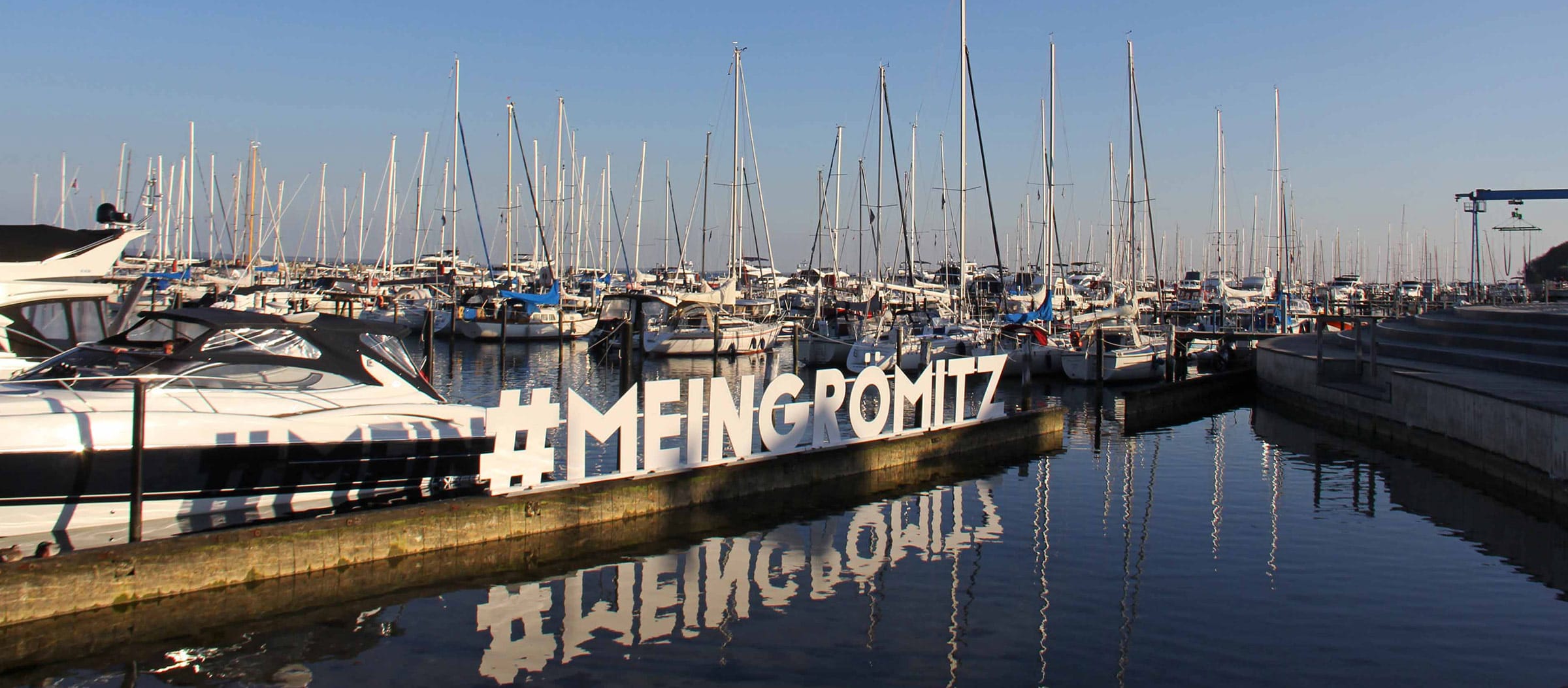 Blick auf den Yachthafen von Grömitz bei Sonnenschein mit zahlreichen Segelbooten und einem großen weißen Schriftzug '#MEINGRÖMITZ' am Kai, der sich im ruhigen Wasser spiegelt.