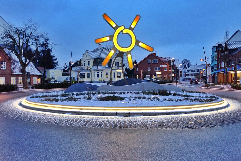 Ein beleuchteter Kreisverkehr in Grömitz bei Dämmerung im Winter, in dessen Mitte eine große, gelbe Metallskulptur in Form einer Sonne steht. Die Umgebung ist leicht mit Schnee bedeckt und im Hintergrund sind typische Wohnhäuser des Küstenortes zu sehen.